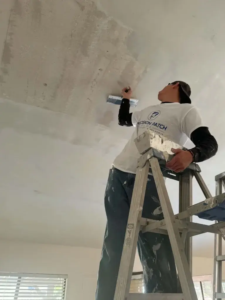 Drywall finisher in a Precision Patch branded shirt skim-coating a ceiling from a ladder
