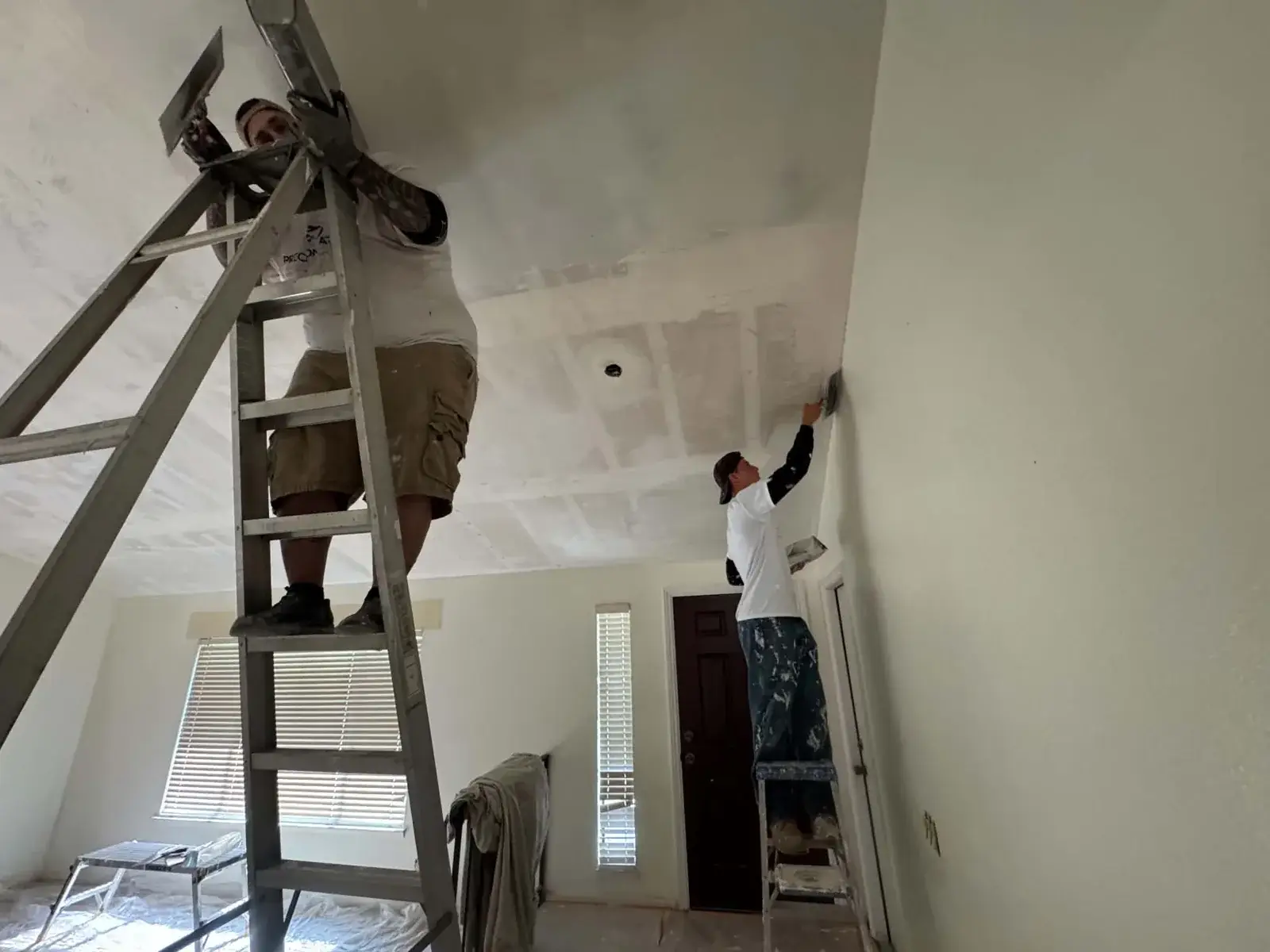 Two drywall finishers on ladders coating a ceiling with joint compound